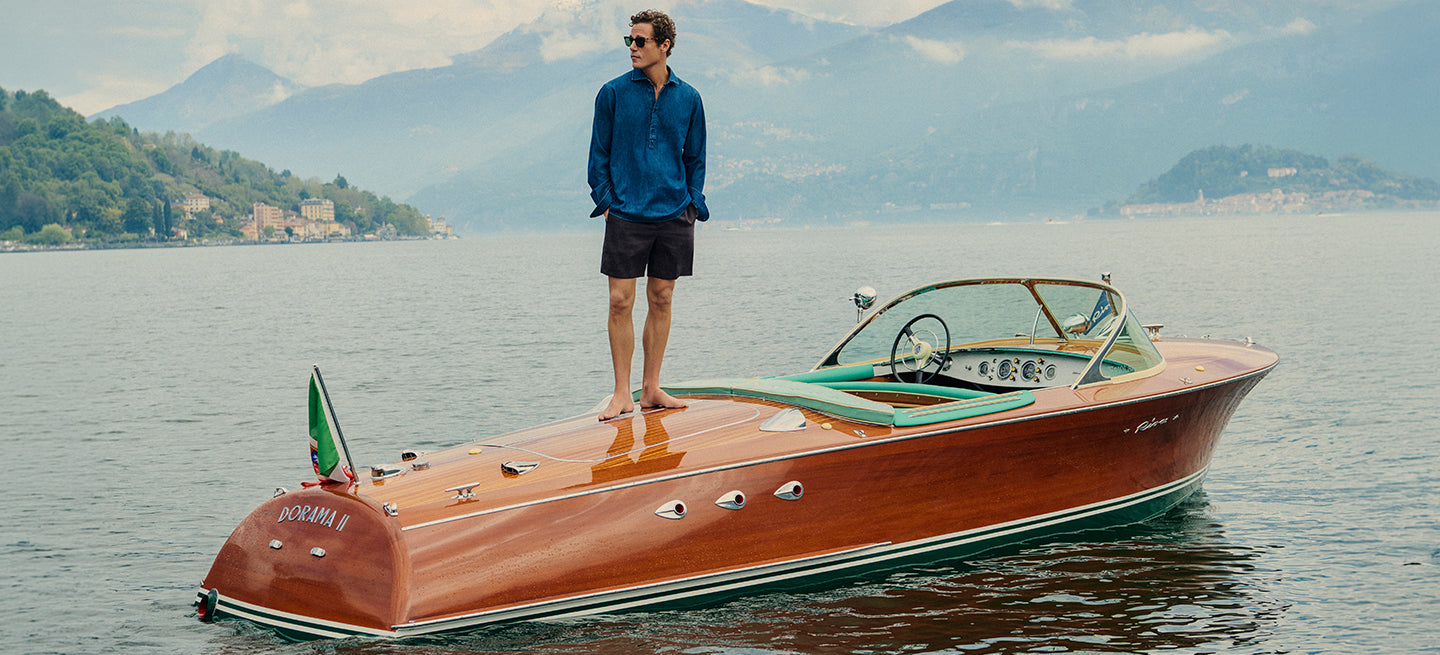 Man standing on a classic wooden boat on a lake with mountains in the background