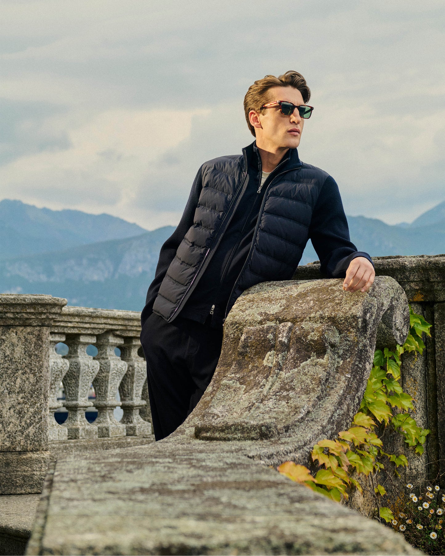 Man in a black puffer jacket and sunglasses leaning on a stone railing with mountains in the background