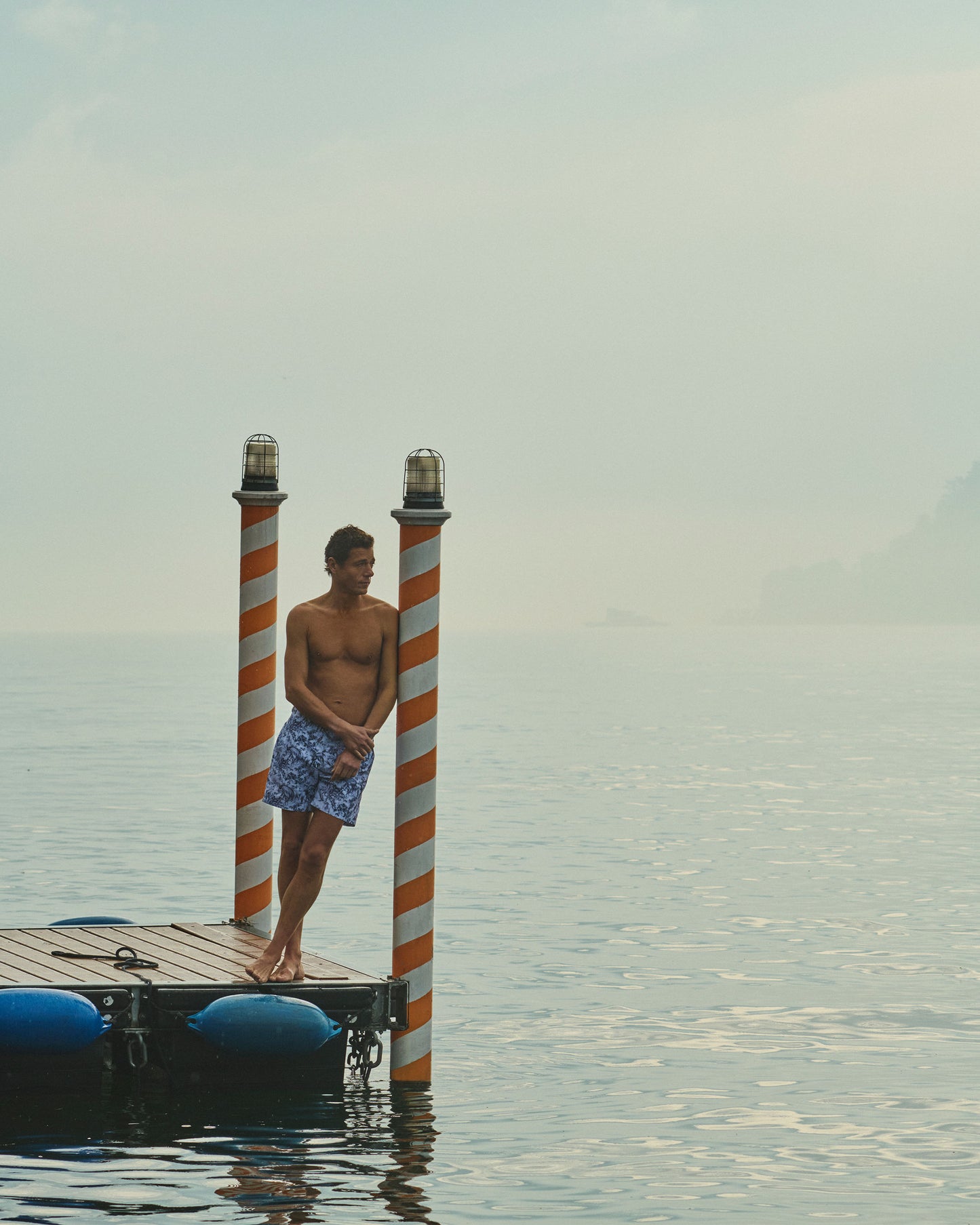 Man standing on a dock with striped posts, surrounded by water and sky.