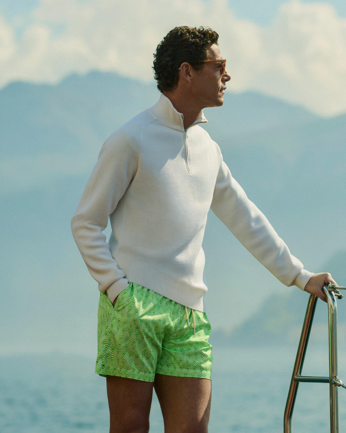 A man wearing a white quarter-zip pullover sweater and bright green patterned swim trunks stands on a boat with water and mountains in the background.
