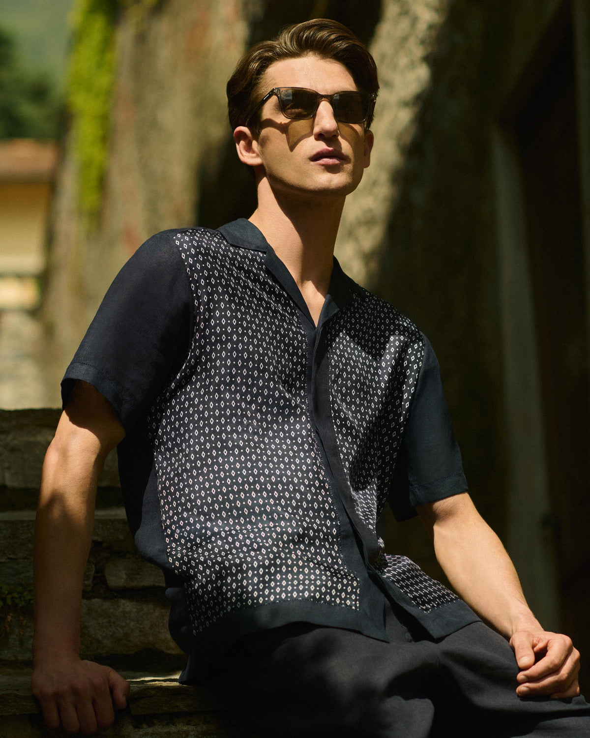 A man wearing dark sunglasses, a navy blue short-sleeve button-up shirt with a white geometric patterned chest panel, and dark trousers, sitting outdoors on stone steps.