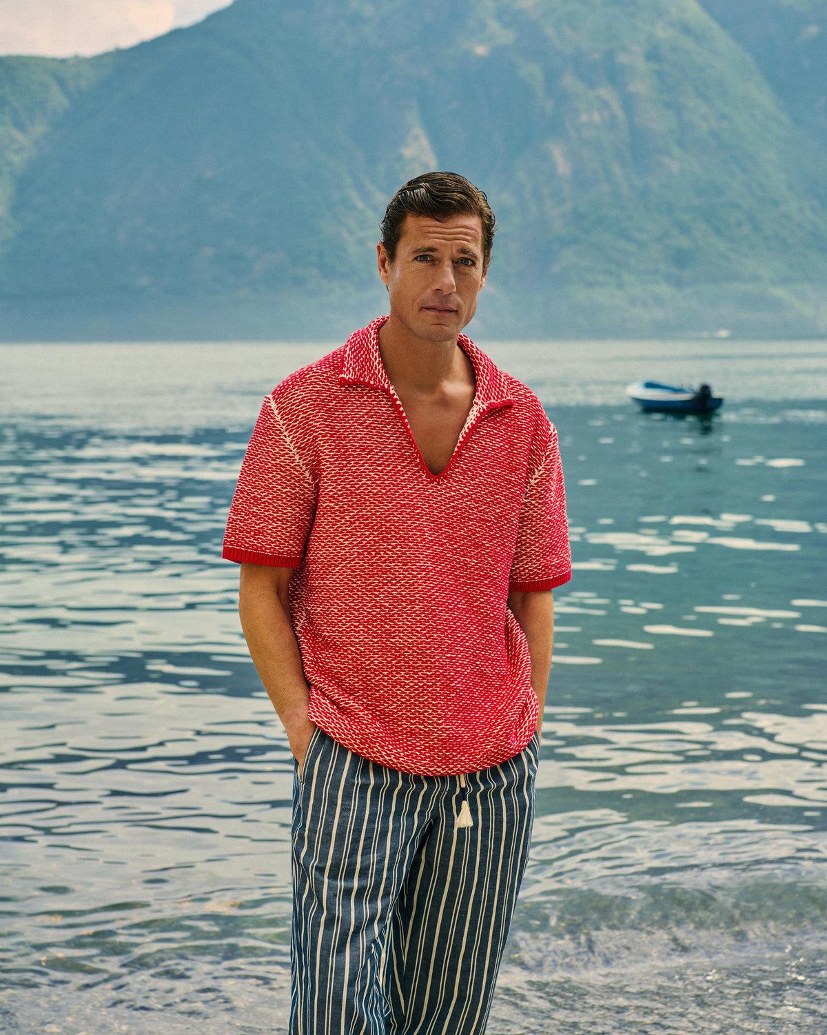 A man standing at the edge of a lake or sea, wearing a red textured short-sleeve polo shirt and blue-and-white vertically striped pants, with mountains and water in the background.
