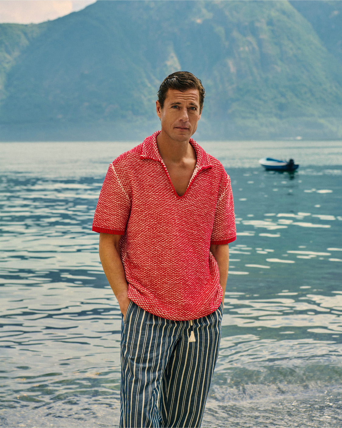 Man wearing red crochet stitch polo shirt with striped trousers against lake background