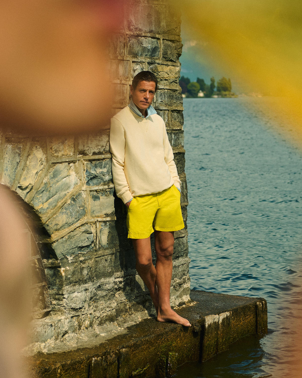 A man stands barefoot on a stone pier by the water, wearing a cream knit sweater over a light blue collared shirt and bright yellow shorts.