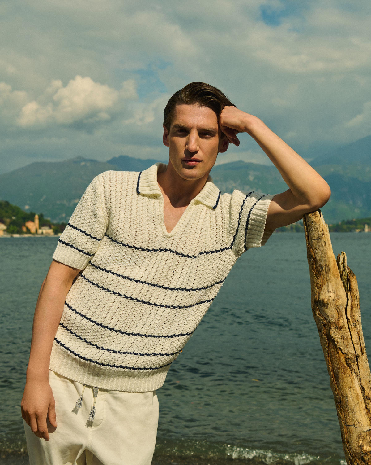 A man wearing a cream knit polo sweater with thin black horizontal stripes and cream drawstring pants, leaning against a driftwood post by a lake with mountains in the background.