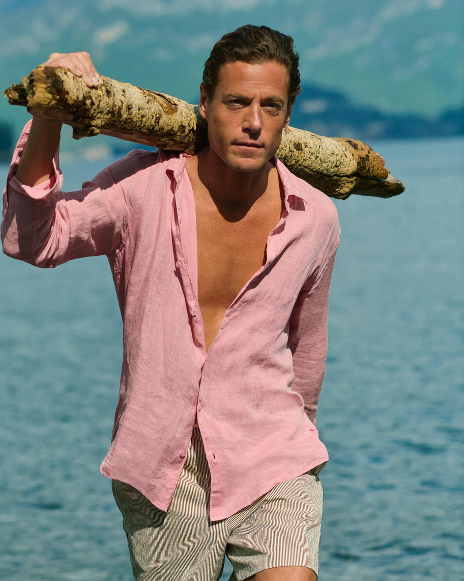 A man standing by a body of water with mountains in the background, wearing an open light pink linen shirt and light beige shorts, carrying a large piece of driftwood over his shoulder.