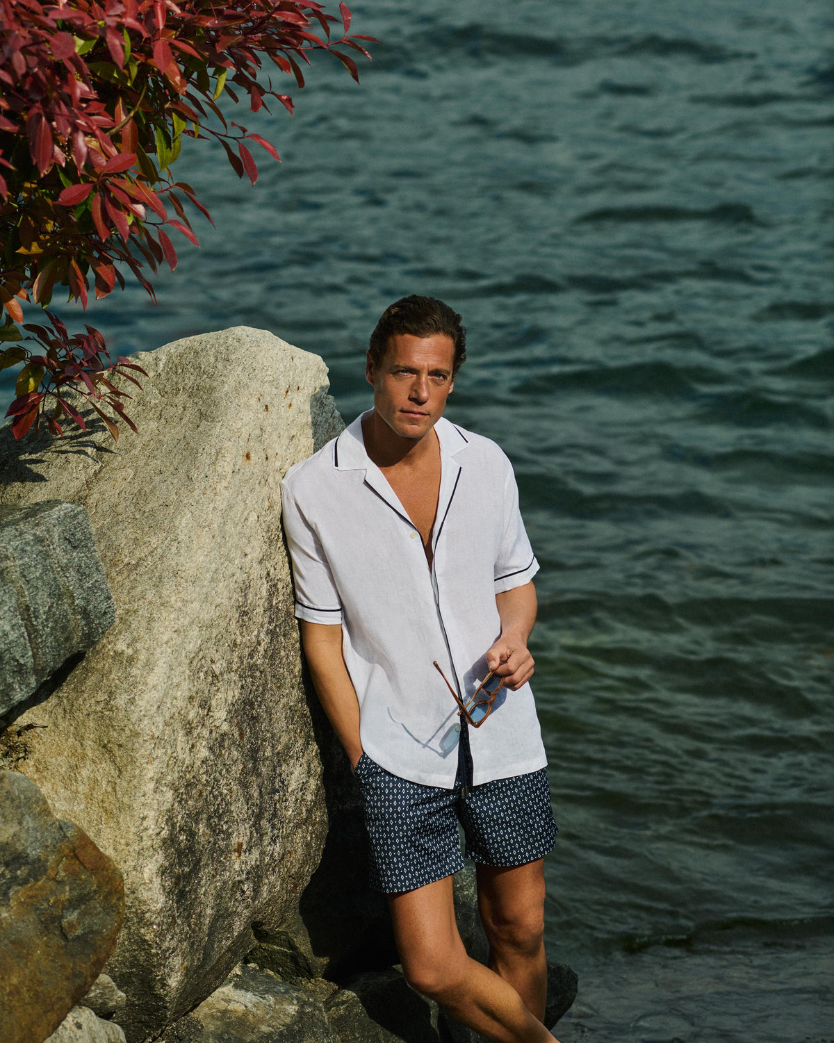 A man stands on rocks by the water wearing a white short-sleeve button-up shirt with dark piping and dark patterned shorts, holding sunglasses.