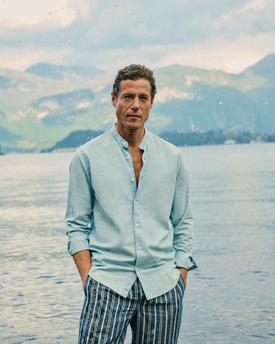 A man standing outdoors by a lake with mountains in the background, wearing a light blue button-up shirt and striped blue-and-white trousers.