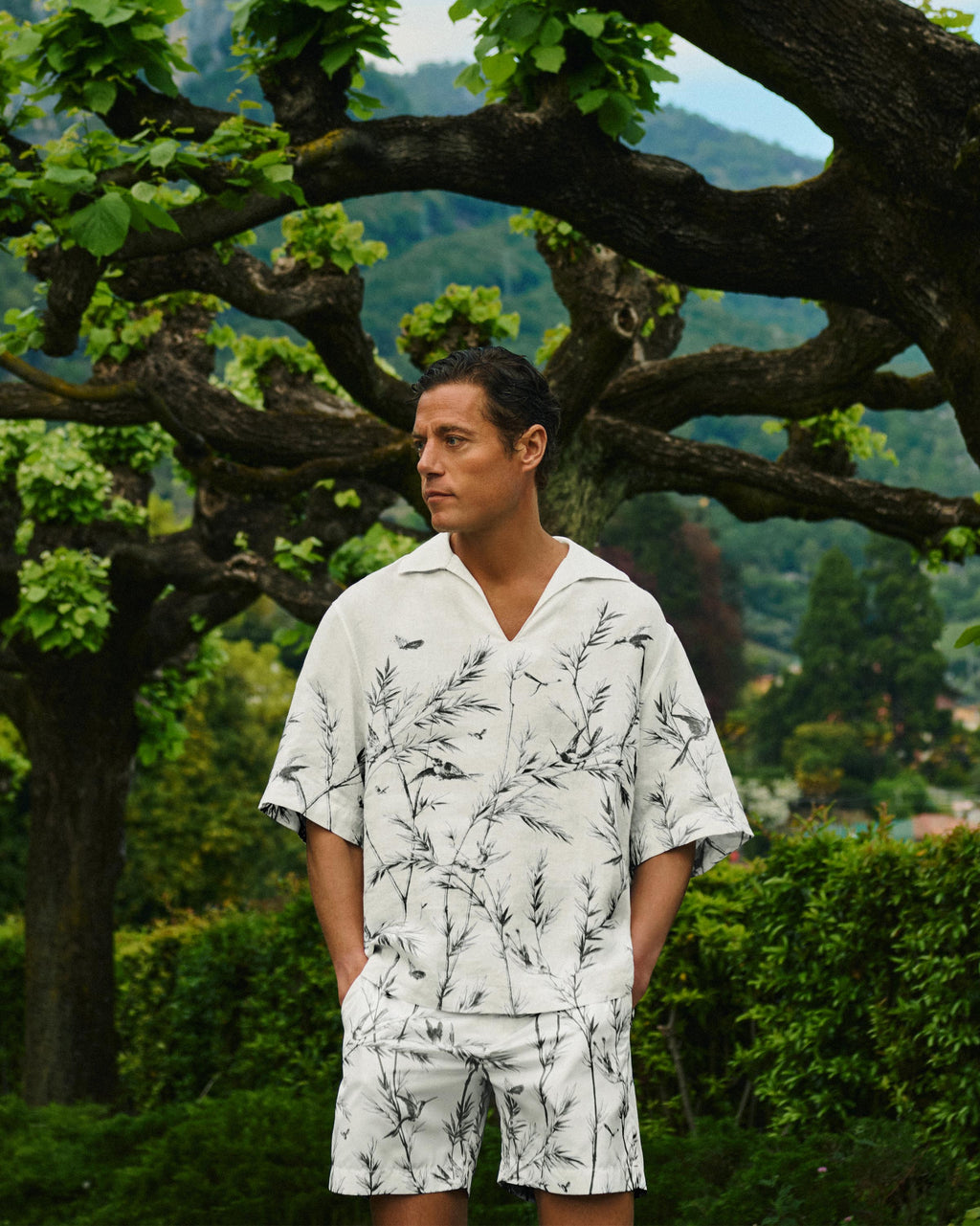 A man wearing a white short-sleeve camp collar shirt and matching shorts with a black botanical leaf print, standing outdoors in front of a large tree with green foliage.
