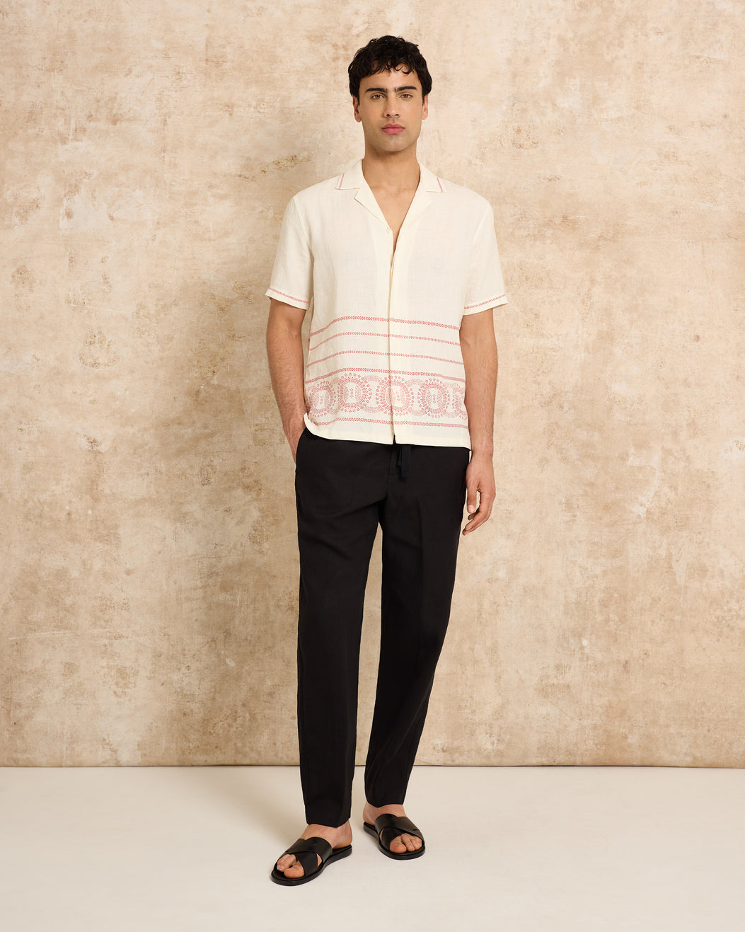 Man wearing a white shirt with red patterns and black pants against a beige wall.