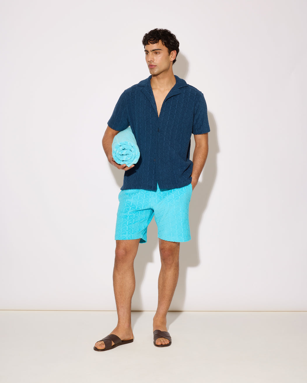 Man wearing a blue shirt and light blue shorts holding a blue towel on a white background