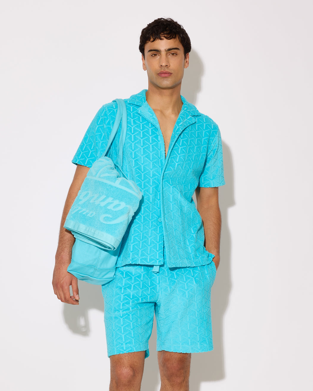 Man wearing a turquoise shirt and shorts with a tote bag and beach towel on a white background