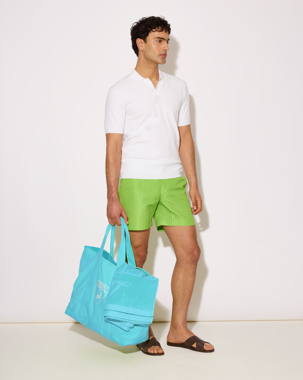 Man holding a turquoise bag wearing a white shirt and green shorts on a white background