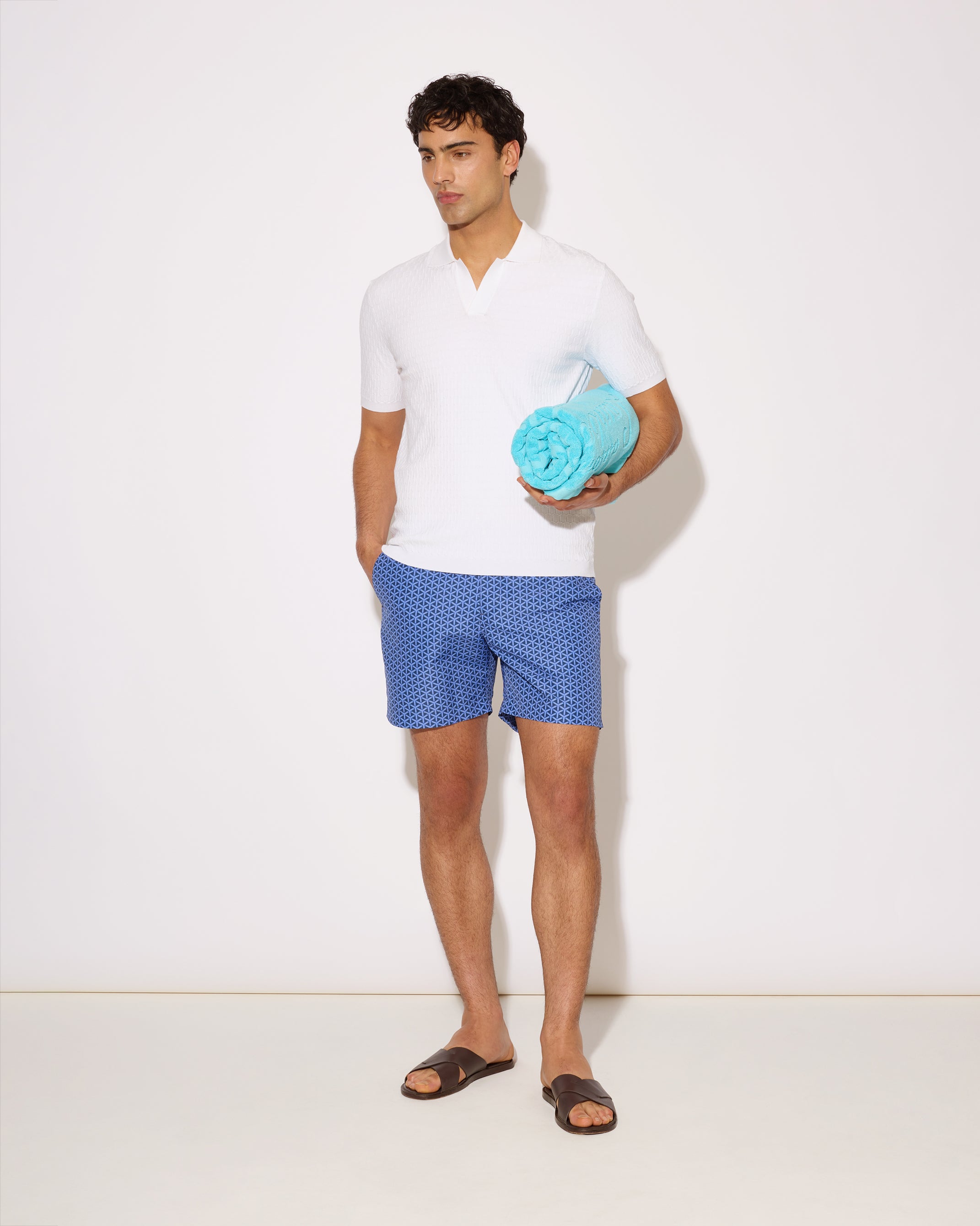 Man holding a blue towel wearing a white shirt and blue patterned shorts on a white background