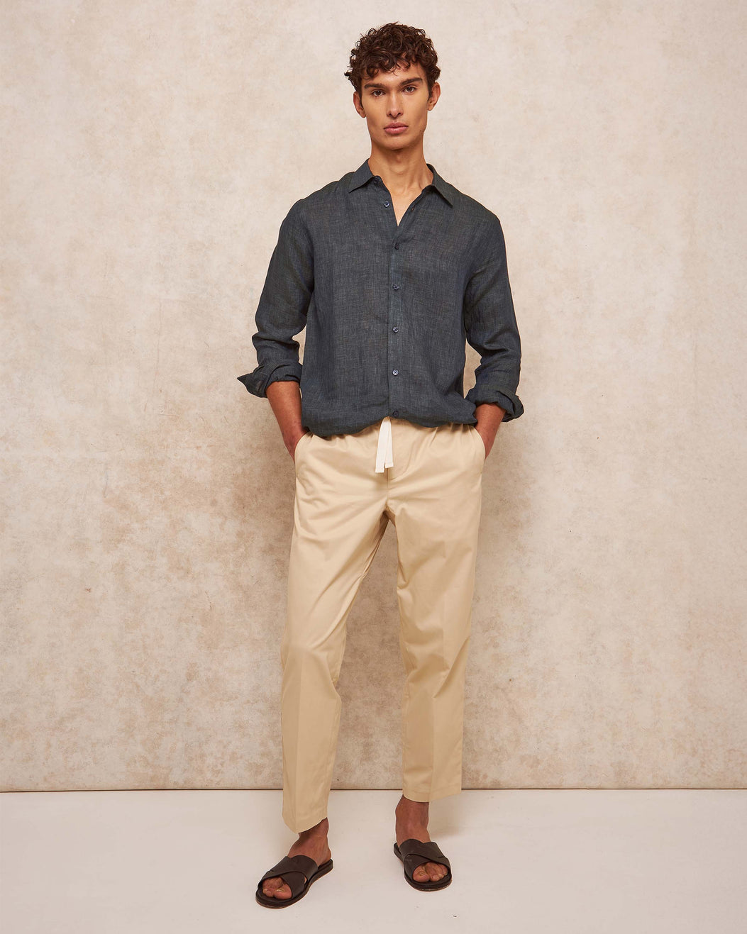 Man wearing a dark denim shirt and beige pants against a beige wall.