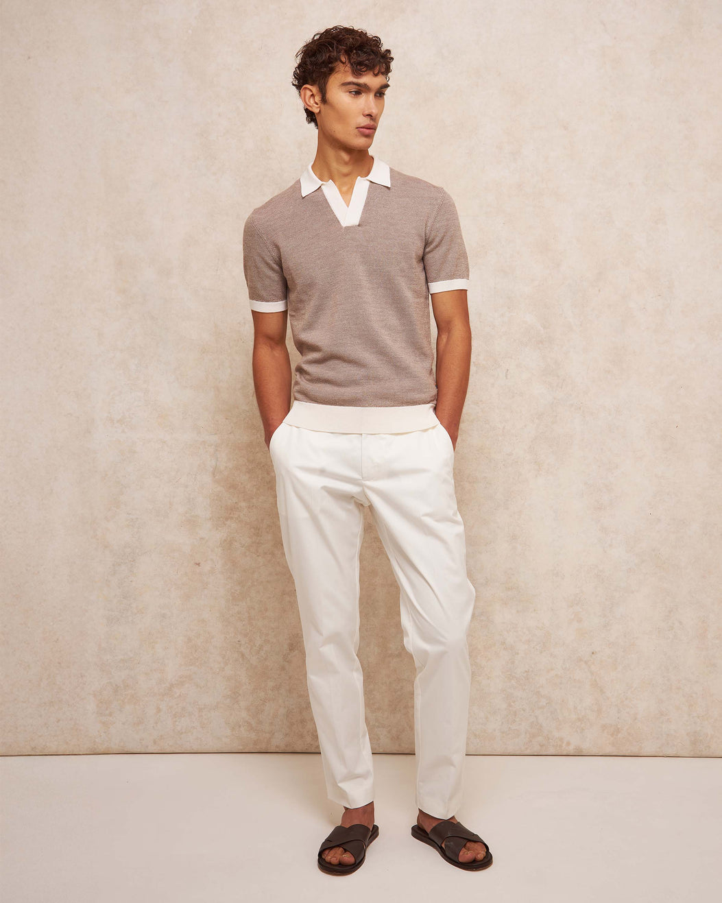Man wearing a gray sweater with white trim and white pants against a beige background