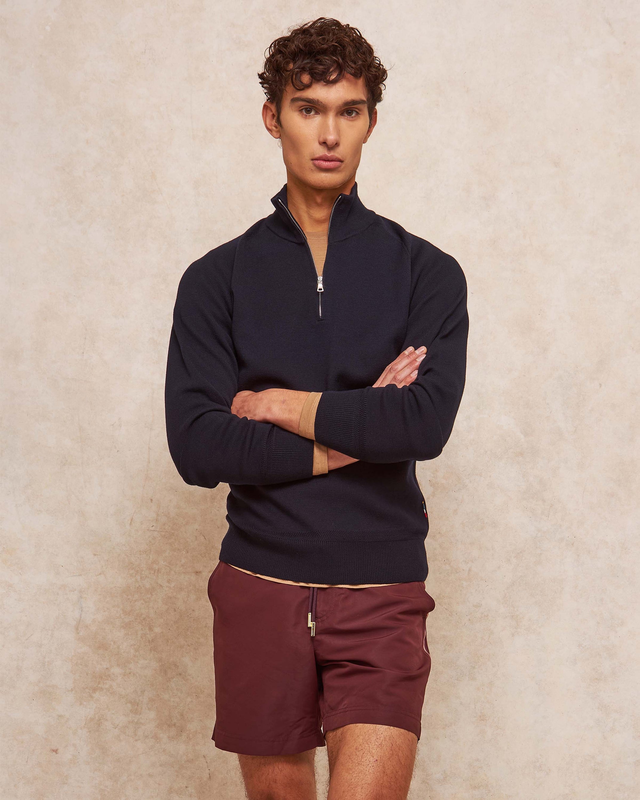 Man wearing a dark blue quarter-zip sweater and maroon shorts against a beige background
