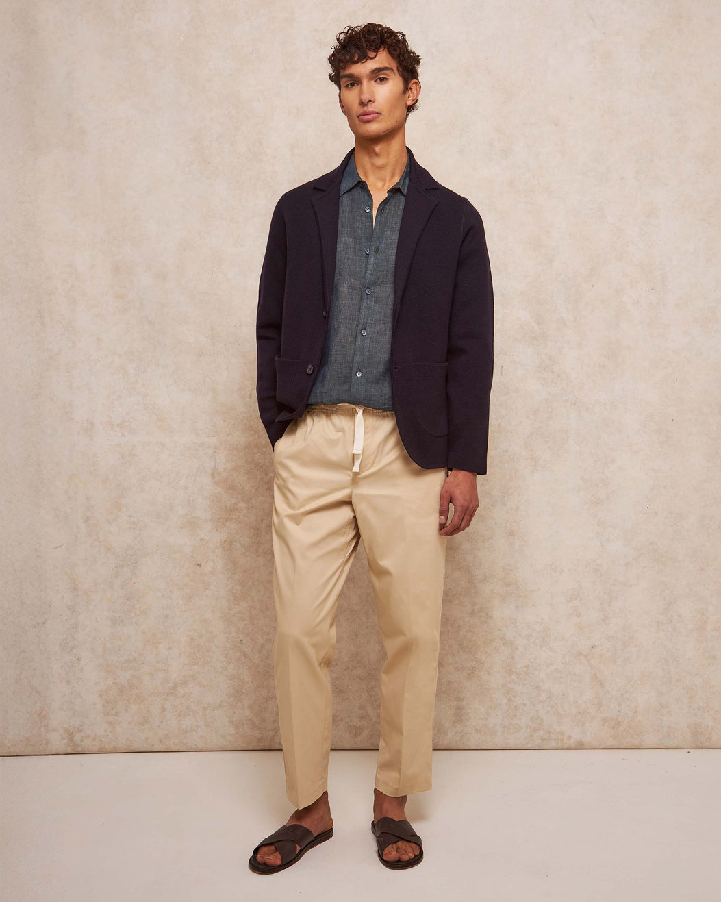 Man wearing a navy blazer, blue shirt, beige pants, and sandals against a neutral background