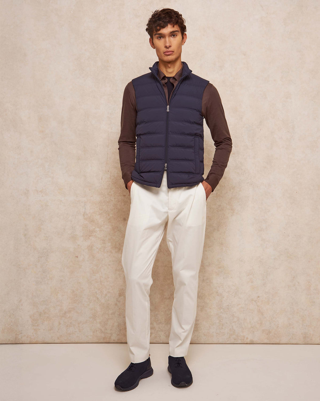 Man wearing a navy puffer vest over a brown shirt with white pants against a beige background
