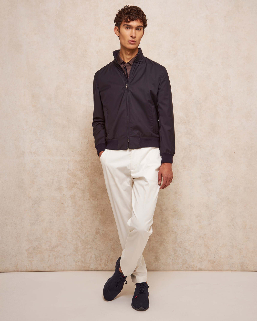 Man wearing a dark jacket, white pants, and black shoes against a beige wall.