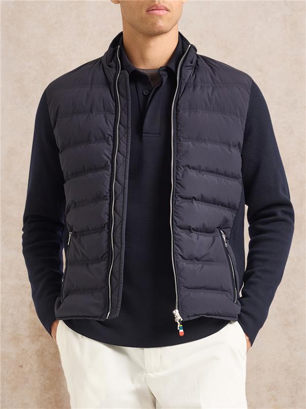 man wearing navy padded jacket with white trousers on a plain background