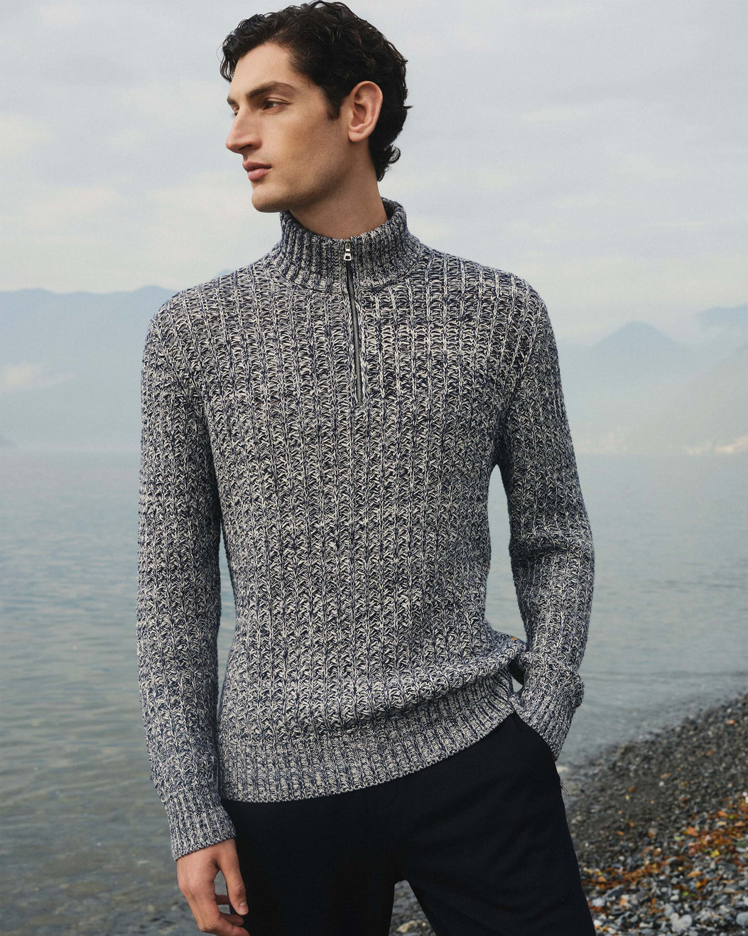 Man wearing a gray knitted sweater standing by a body of water with mountains in the background