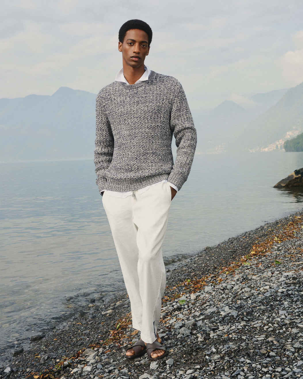 Man standing on a pebbly beach with mountains in the background