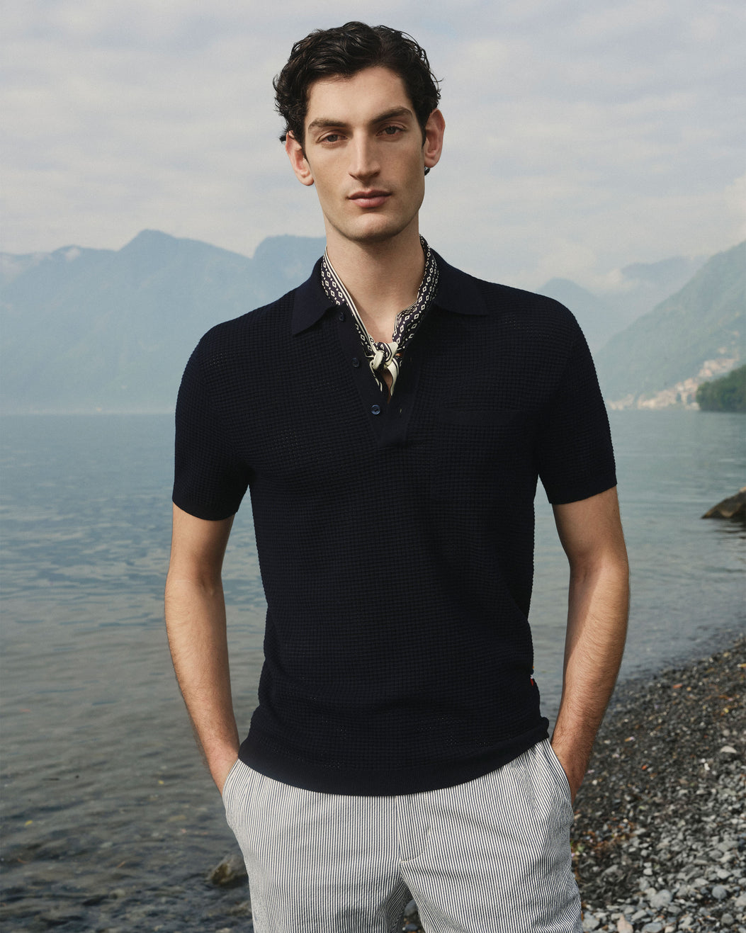 A man wearing a dark navy short-sleeve knit polo shirt and light gray trousers, standing on a pebble shore with water and mountains in the background.