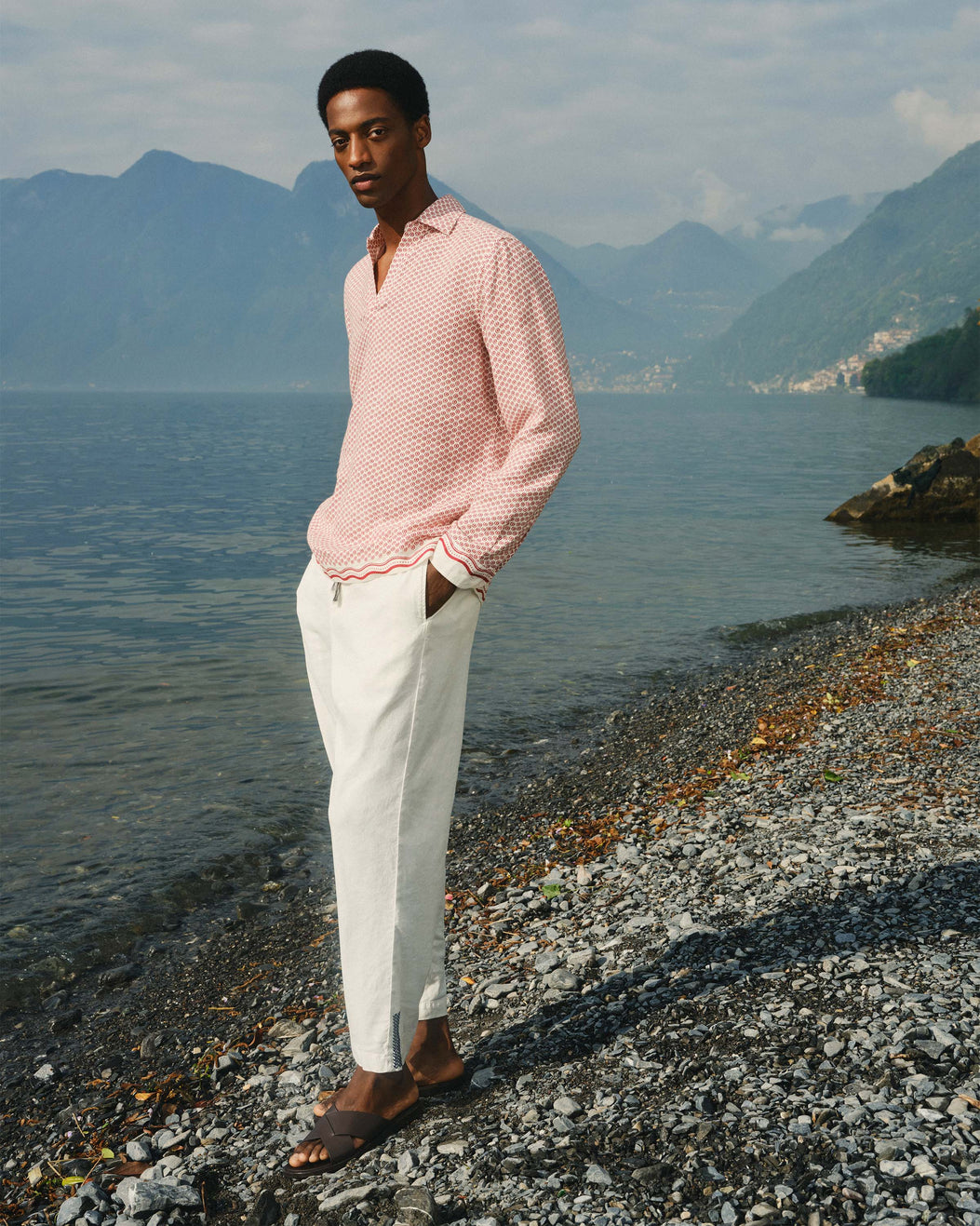Man in a pink shirt and white pants standing on a rocky shore with mountains in the background