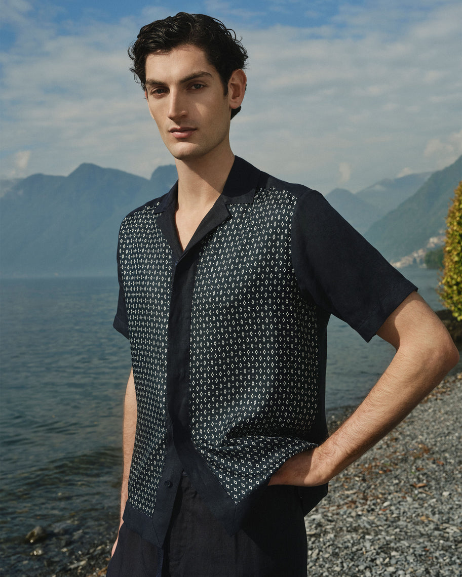 A man wearing a short-sleeve button-up shirt with a dark navy base and a white geometric pattern across the front panels, photographed outdoors by the water with mountains in the background.