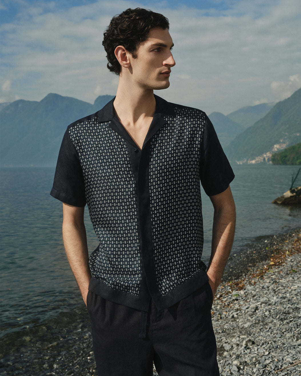 A man wearing a short-sleeve button-front shirt with a dark navy base and a dense small white geometric pattern across the body, photographed outdoors by a lake with mountains in the background.
