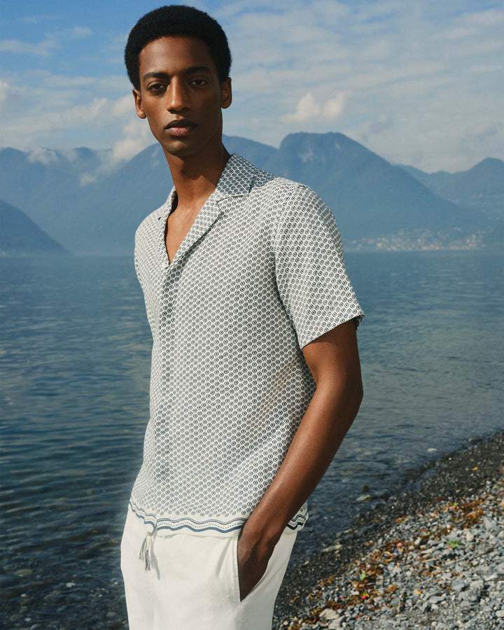 Man standing on a pebbly beach with mountains and water in the background