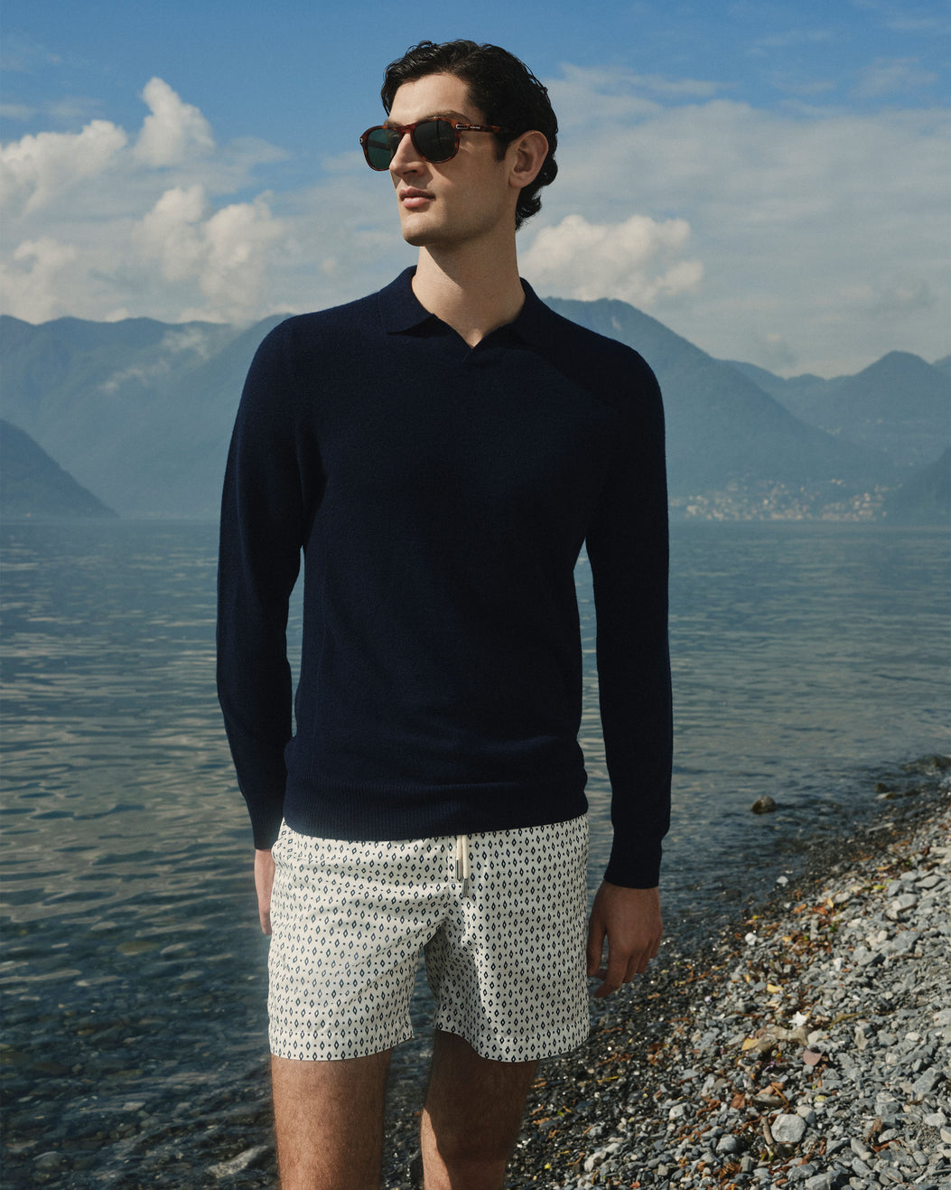 A man wearing a dark navy long-sleeve polo shirt and white shorts with a small black pattern stands on a pebble shore with a lake and mountains in the background.