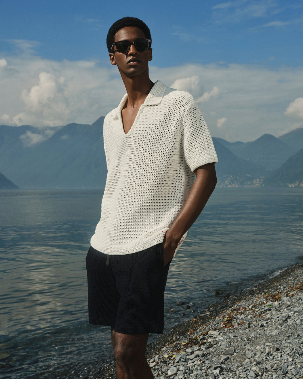 A man wearing a white open-knit short-sleeve polo shirt and dark shorts stands on a rocky lakeshore with mountains and blue sky in the background.