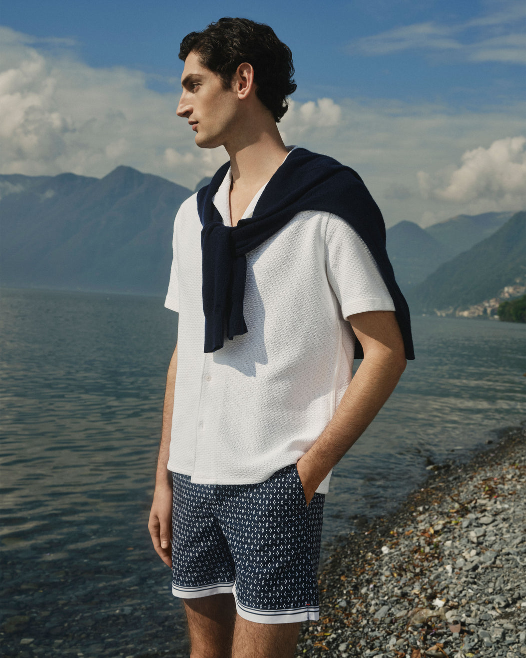A man stands on a pebble shore beside a lake with mountains in the background, wearing a white short-sleeve textured shirt, navy patterned shorts, and a navy sweater draped over his shoulders.