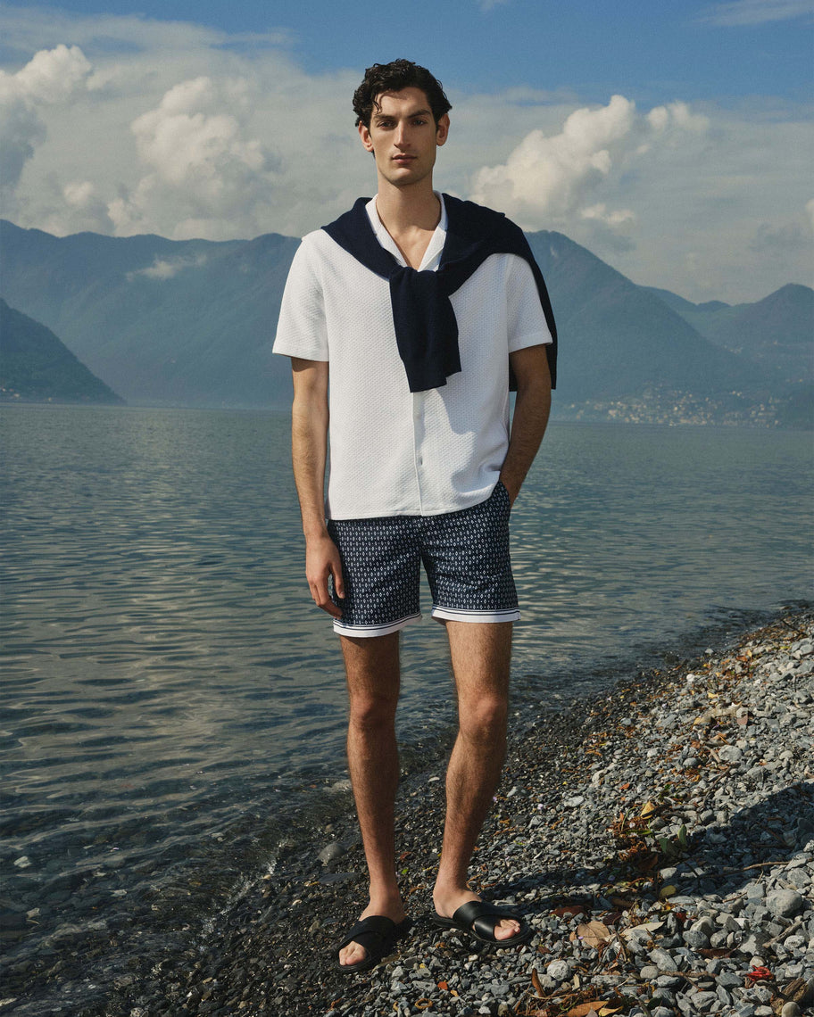 A man stands on a pebble shoreline by a lake with mountains and a cloudy sky in the background. He is wearing a white short-sleeve button-up shirt with a dark navy neck scarf draped over his shoulders, navy shorts with a small white geometric pattern, and black sandals.