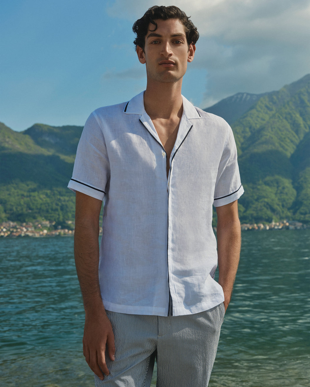 A man wearing a white short-sleeve linen camp collar shirt with dark piping trim, standing outdoors in front of a scenic lake and green mountains.