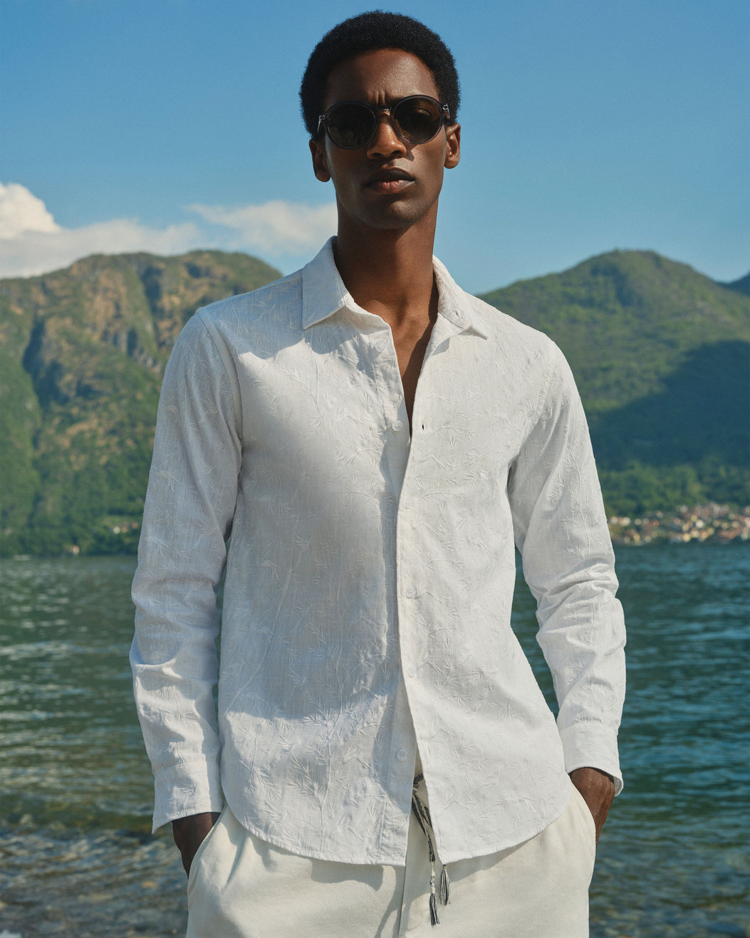 A man wearing a white textured long-sleeve button-up shirt and white drawstring pants, standing outdoors in front of a scenic lake and mountain background.