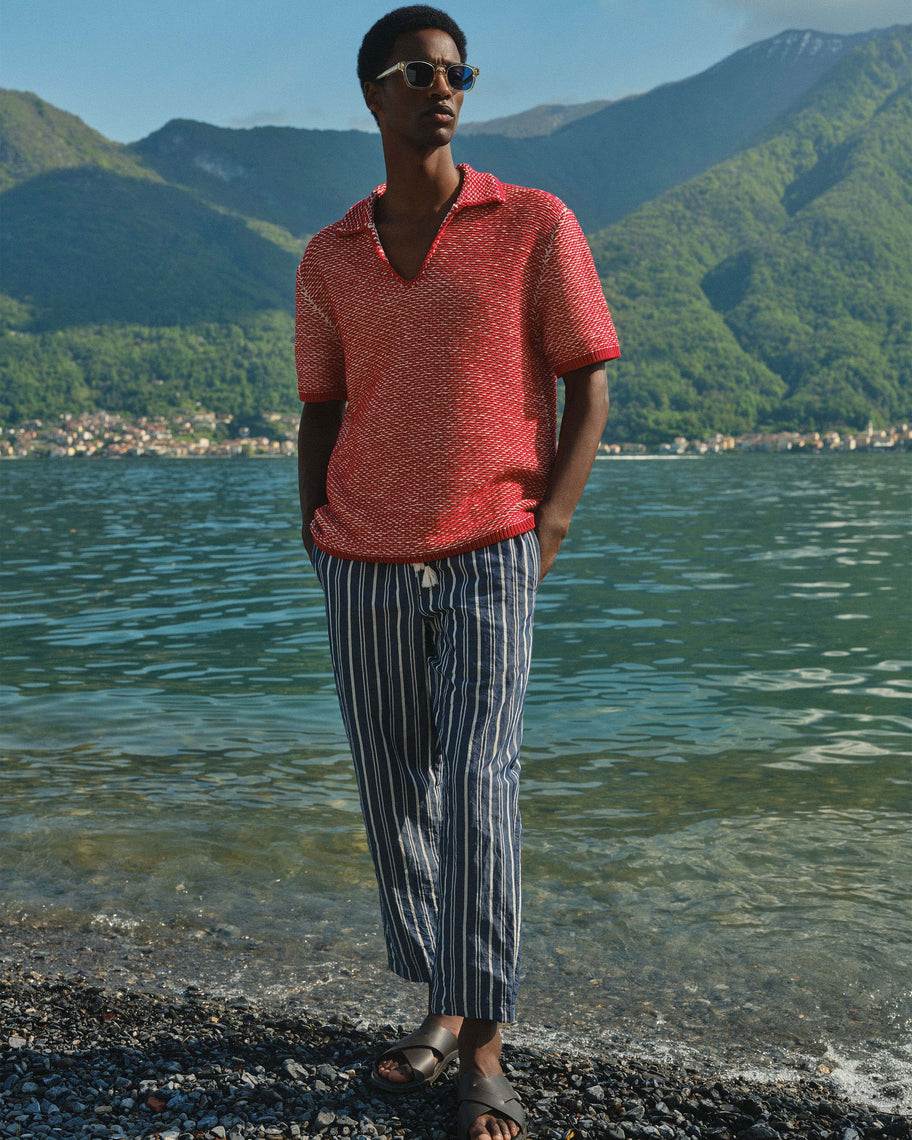 A man wearing a textured coral-red short-sleeve polo shirt and blue-and-white vertically striped trousers stands on a pebble shore with a lake and green mountains in the background.