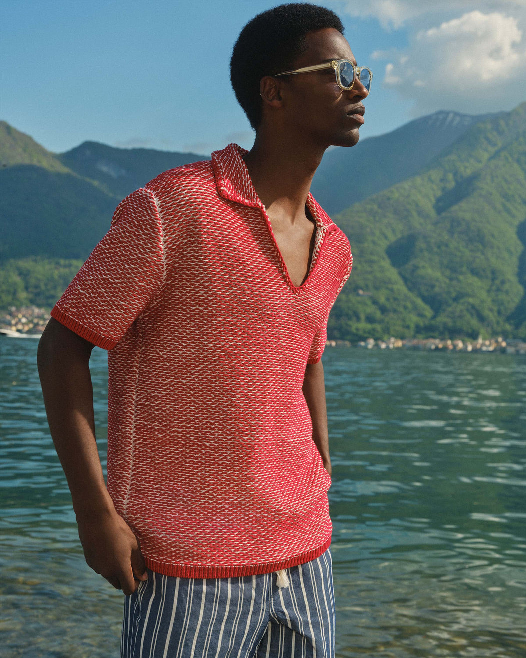 A man wearing a coral-red and white textured knit short-sleeve polo shirt and striped shorts, standing by a lake with mountains in the background.