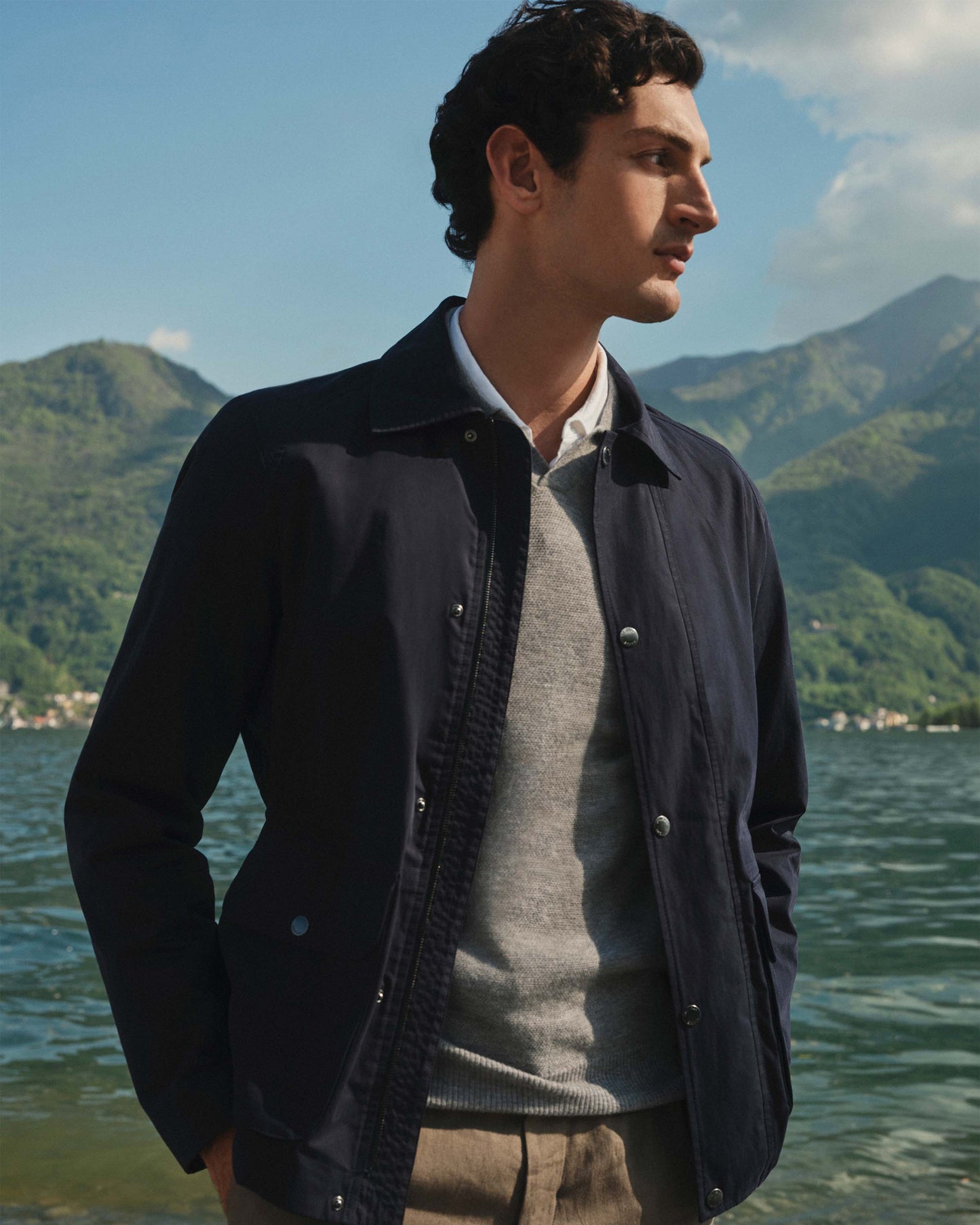 Man wearing a dark jacket over a gray sweater by a lake with mountains in the background