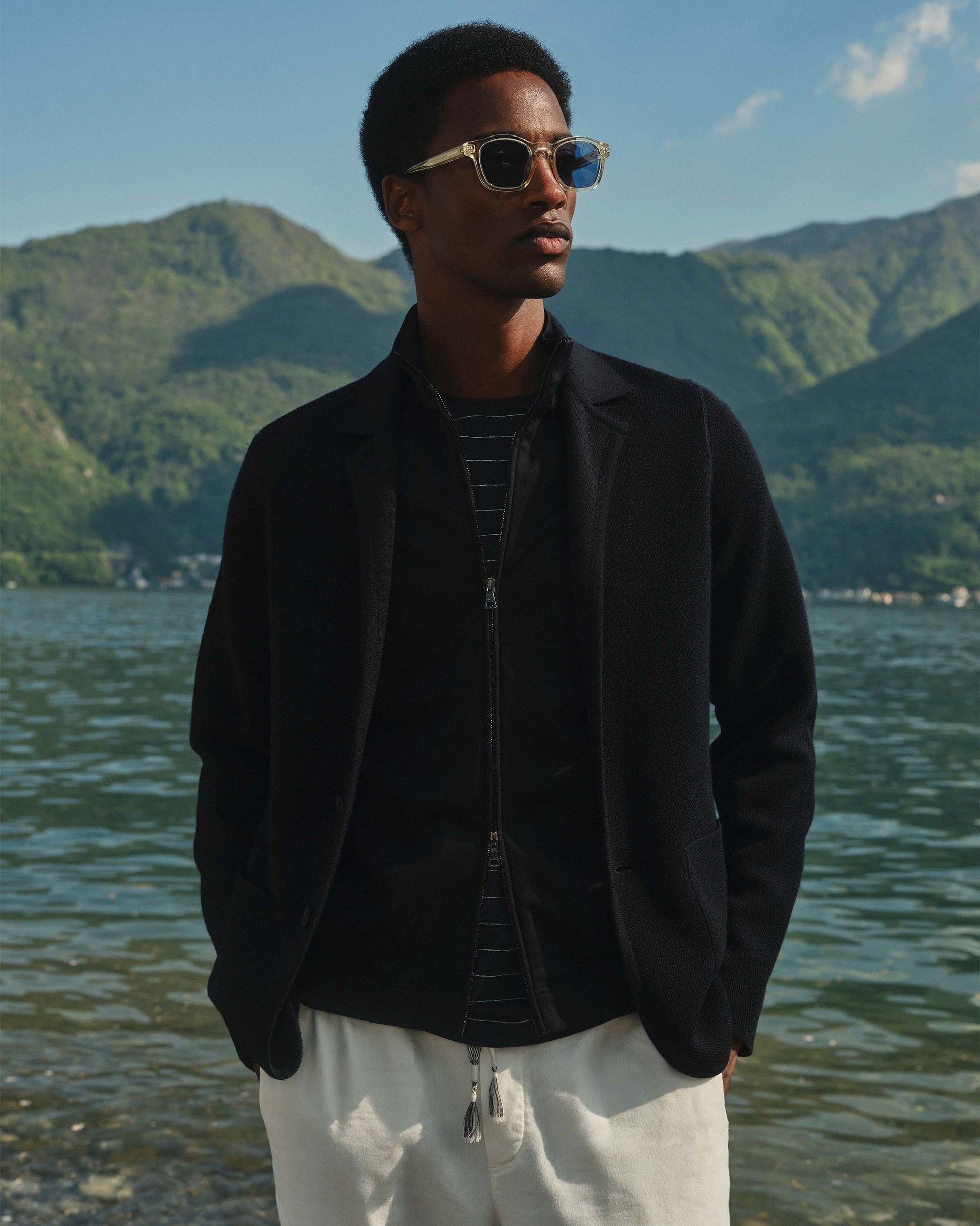 Man wearing sunglasses and a black jacket standing by a lake with mountains in the background