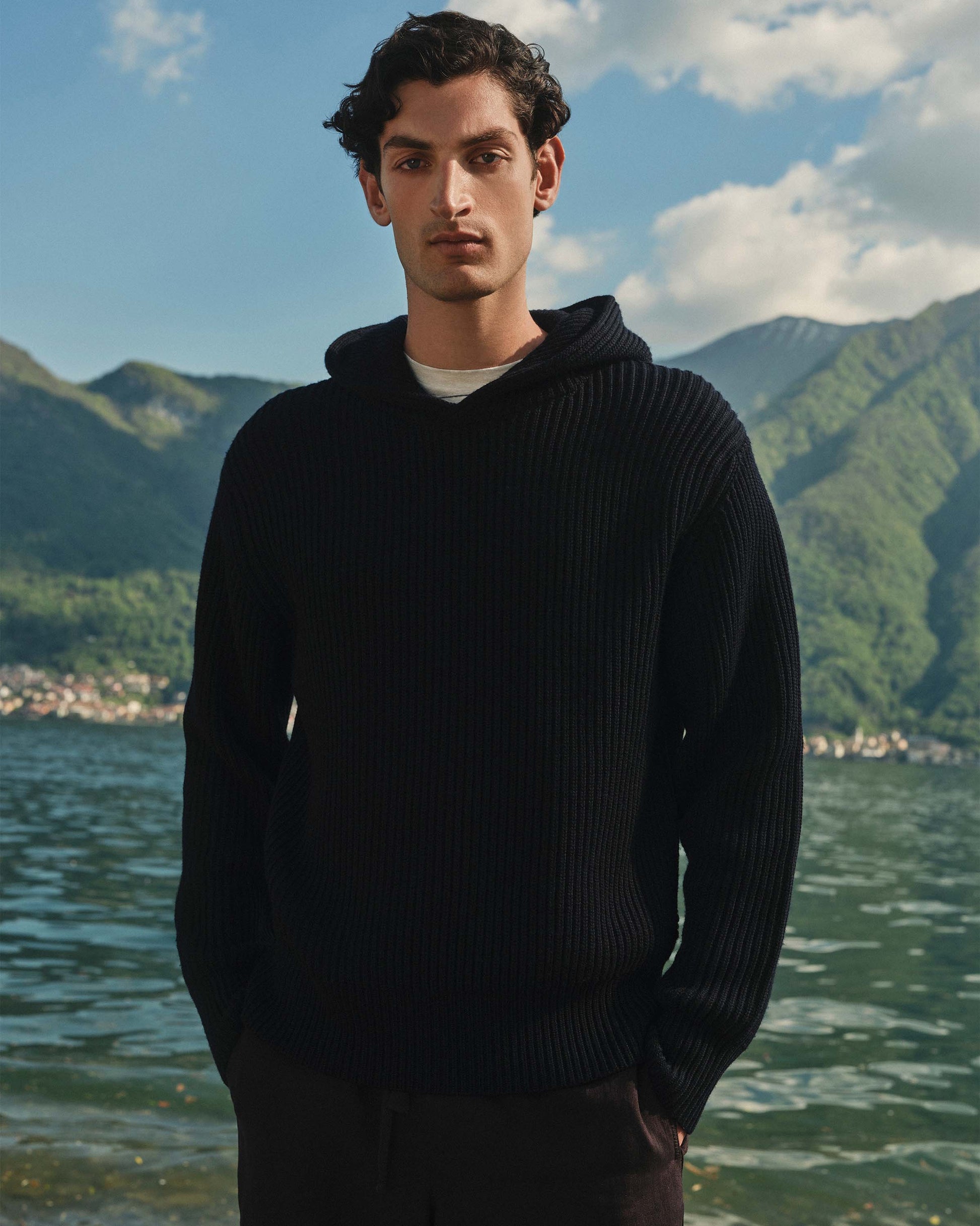 Man wearing a black sweater standing by a lake with mountains in the background