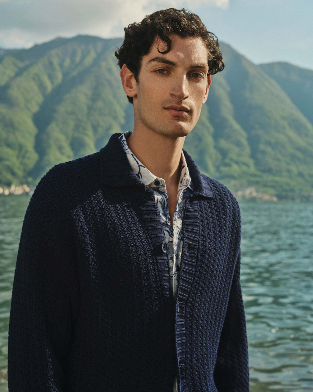 Man wearing a dark blue knitted cardigan over a patterned shirt with a mountainous landscape in the background.