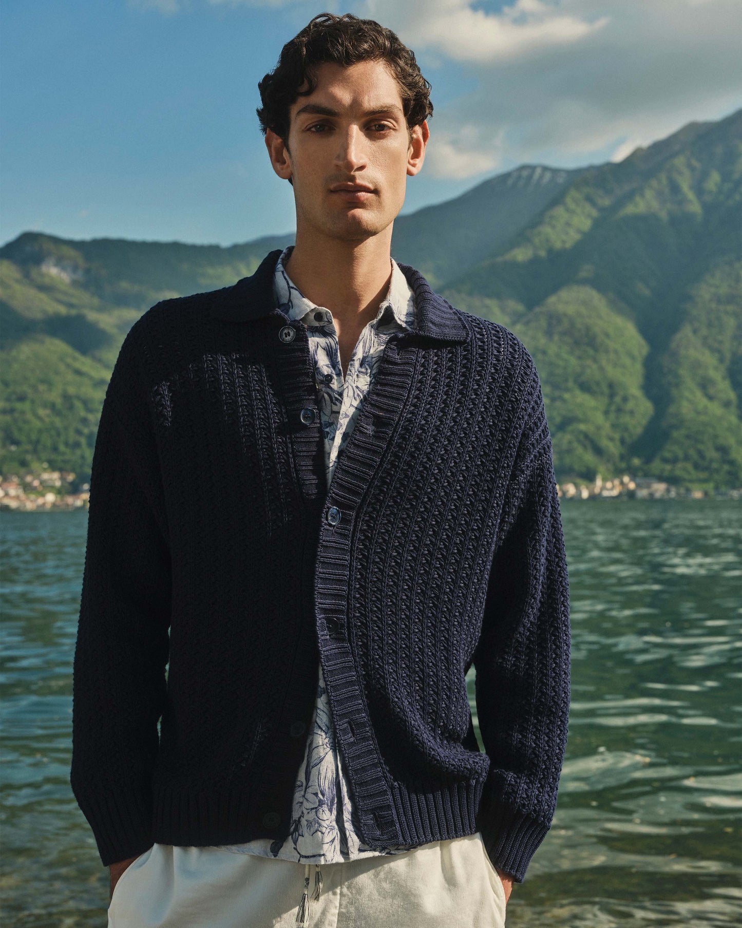 Man wearing a dark blue knitted cardigan by a lake with mountains in the background