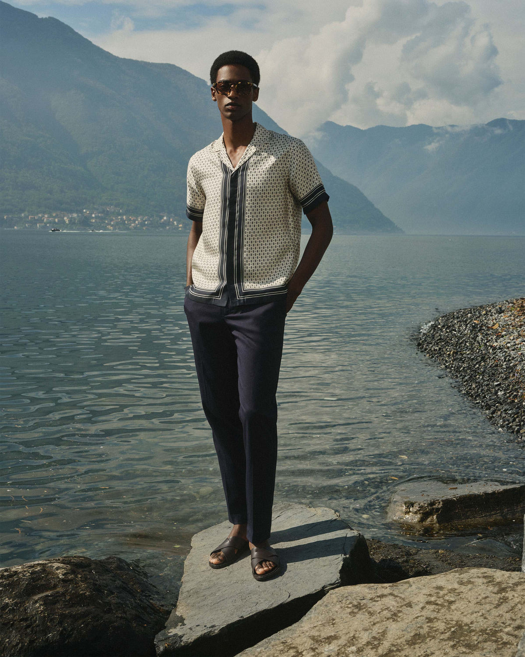 A man wearing a cream short-sleeve button-up shirt with a black vertical stripe and black geometric pattern, paired with dark trousers, sunglasses and brown sandals standing on a rock by a lake with mountains in the background.