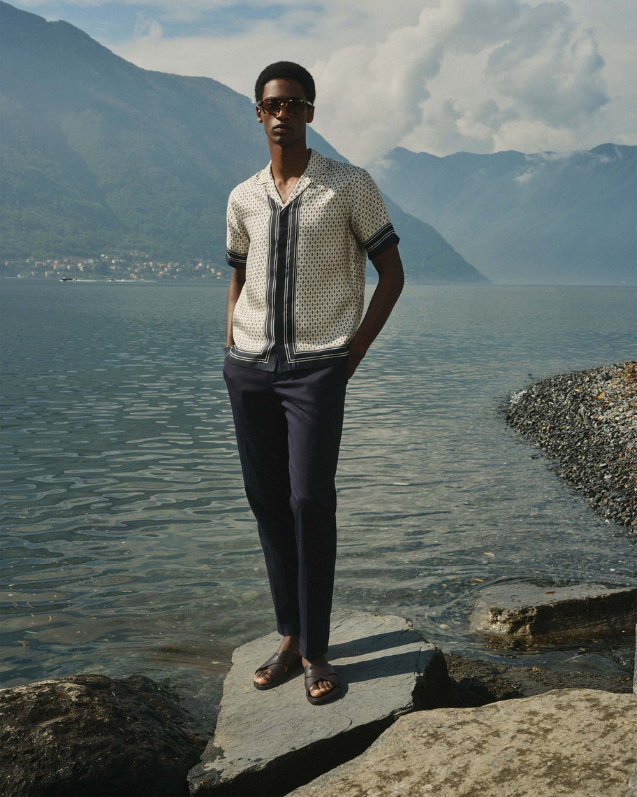 A man wearing a cream short-sleeve button-up shirt with a black vertical stripe and black geometric pattern, paired with dark trousers, sunglasses and brown sandals standing on a rock by a lake with mountains in the background.
