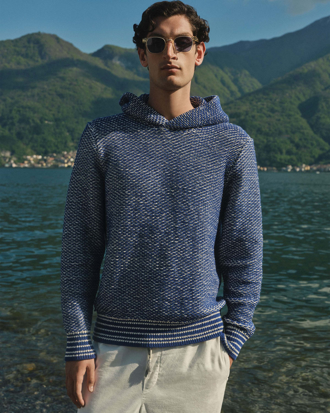 A man wearing a textured navy and white knit hoodie with sunglasses, standing in front of a scenic lake and green mountain background.