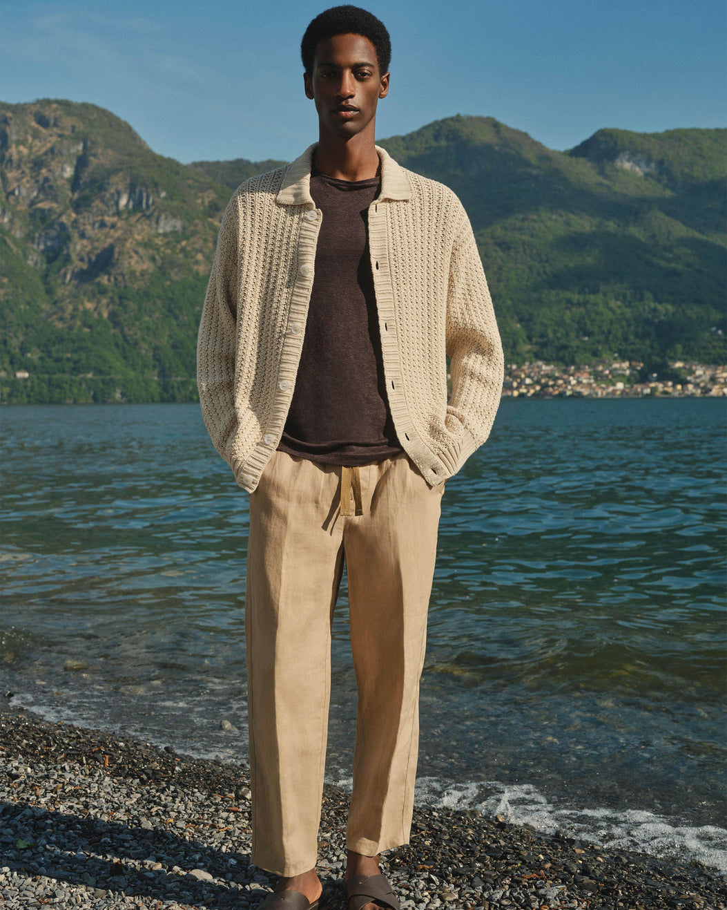 A man stands on a pebble beach with water and mountains in the background, wearing a cream textured cardigan over a dark brown shirt and beige drawstring trousers.
