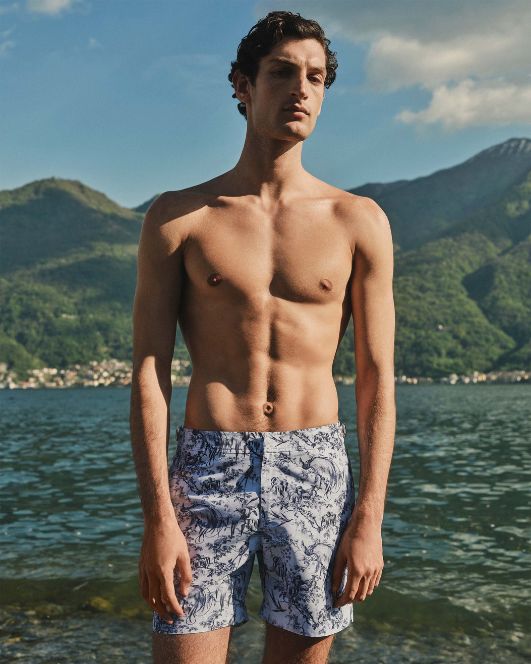Man wearing blue patterned swim shorts standing by a lake with mountains in the background